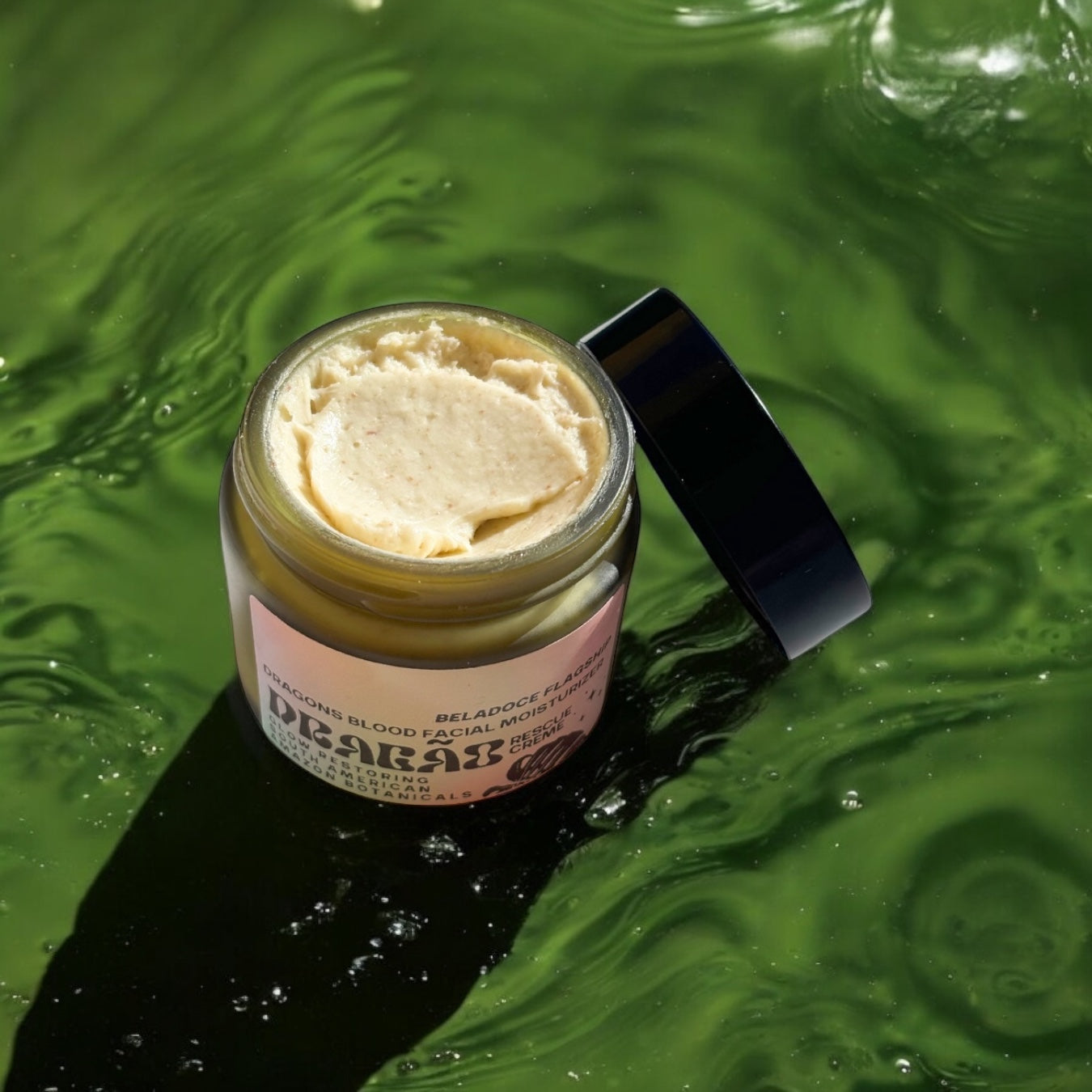 The Beladoce Botanicals Dragão Rescue Creme jar with the lid slightly open, showcasing the creamy, buttery texture of the winter reserve formula. A wooden spatula lies beside it, ready for application. The backdrop includes a hint of greenery, reinforcing its Amazonian origins.